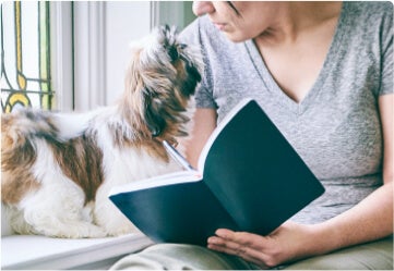 Donna con un libro in mano mentre guarda il cane accanto a lei.